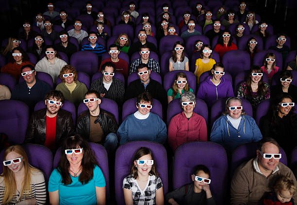 A wide shot of a large audience wearing 3D glasses in a darkened movie theater.